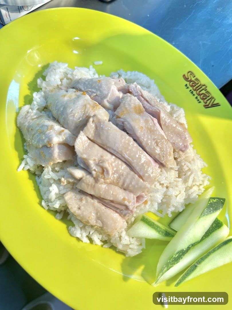 A plate of chicken rice with sliced chicken, rice, and cucumber slices. The plate has a logo that says "Sataay by the bay".
