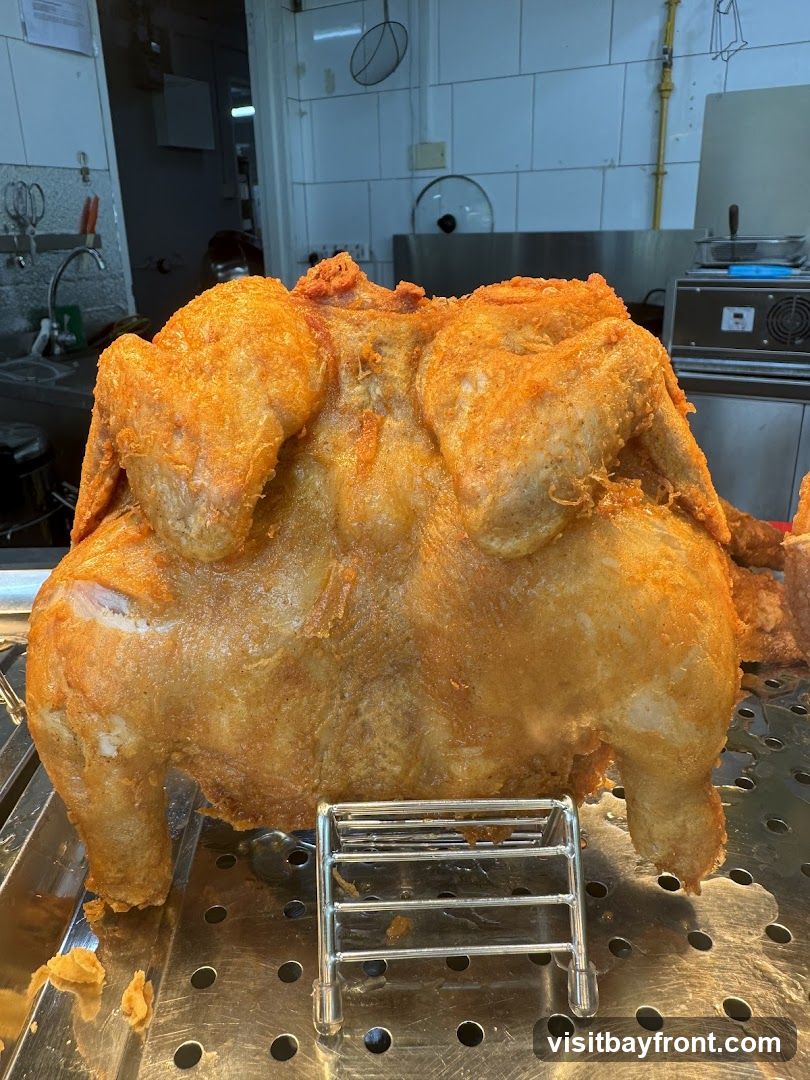 A whole, golden-brown fried chicken is displayed on a metal rack. The chicken appears seasoned with a reddish spice. In the background, a commercial kitchen setting is visible with stainless steel counters, sinks, and equipment.