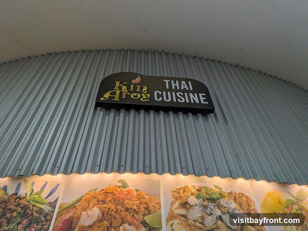 A sign for "Kin Aroy Thai Cuisine" is mounted on a corrugated metal wall. Below the sign are illuminated images of various Thai dishes, including fried rice with shrimp and an omelet with oysters.
