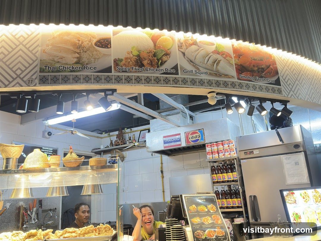 A food stall with images of Thai dishes displayed above. The dishes shown are Thai Chicken Rice, Spicy Fried Chicken Rice, Grill Squid, and Smoked Fresh Crab. Below, staff members are visible, and a menu board displays various food items. Bottles of drinks and cans are stacked on shelves.