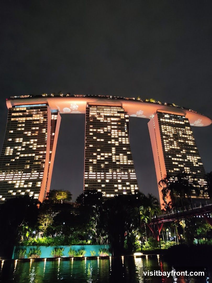A nighttime view of the Marina Bay Sands hotel in Singapore, illuminated with many lights. The iconic building features three towers connected by a large rooftop structure. The rooftop is decorated with illuminated snowflake patterns. Below the hotel, lush greenery and a body of water with reflections are visible. A pedestrian bridge with red supports is also present in the foreground.