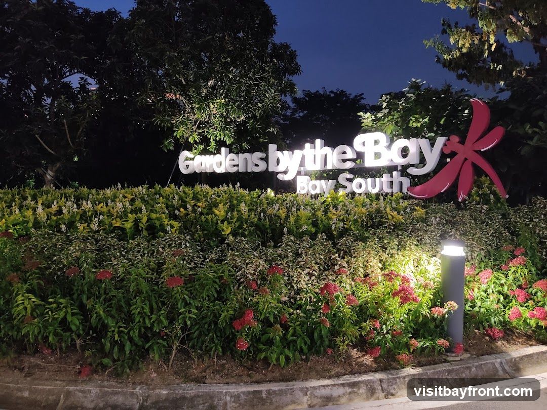 A large, illuminated sign reads "Gardens by the Bay" with "Bay South" below it. To the right of the text is a stylized red dragonfly logo. The sign is set against a backdrop of lush green foliage and flowering plants, illuminated by spotlights. The image is taken at night.