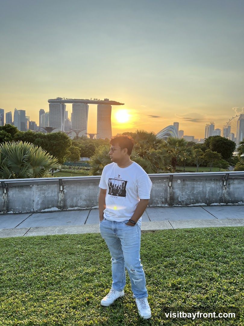 A man stands in a grassy area, looking out at the cityscape of Singapore during sunset. The iconic Marina Bay Sands hotel is prominently visible in the background, along with other skyscrapers and the Singapore Flyer Ferris wheel. The sun is setting, casting a warm glow over the scene. The man is wearing a white t-shirt with a graphic print, light blue ripped jeans, and white sneakers.