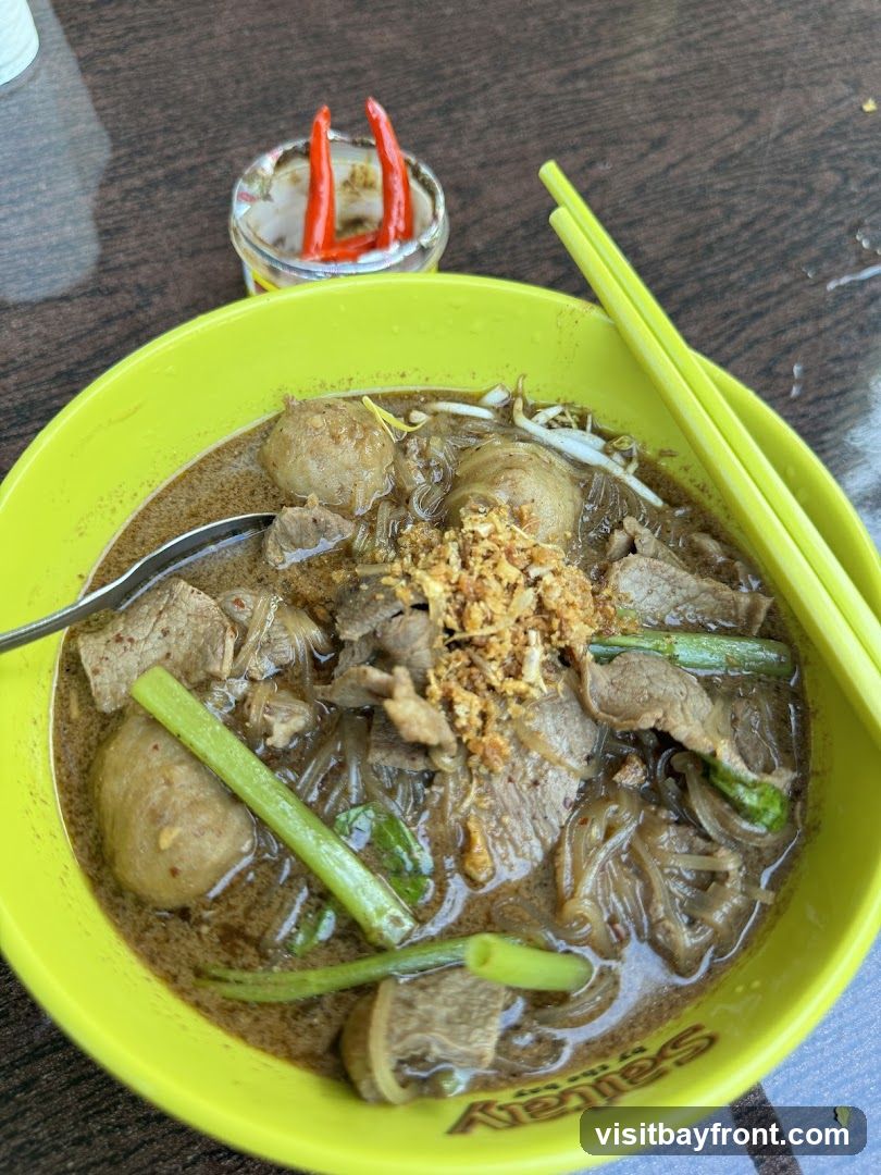 A close-up shot of a bowl of noodle soup. The soup is in a bright yellow bowl and contains noodles, meatballs, sliced beef, bean sprouts, and green onions. Fried garlic is sprinkled on top. A small dish with sliced red chilies in a clear liquid is visible in the background, along with a pair of yellow chopsticks. The bowl has some text on the bottom edge, but it is mostly obscured.