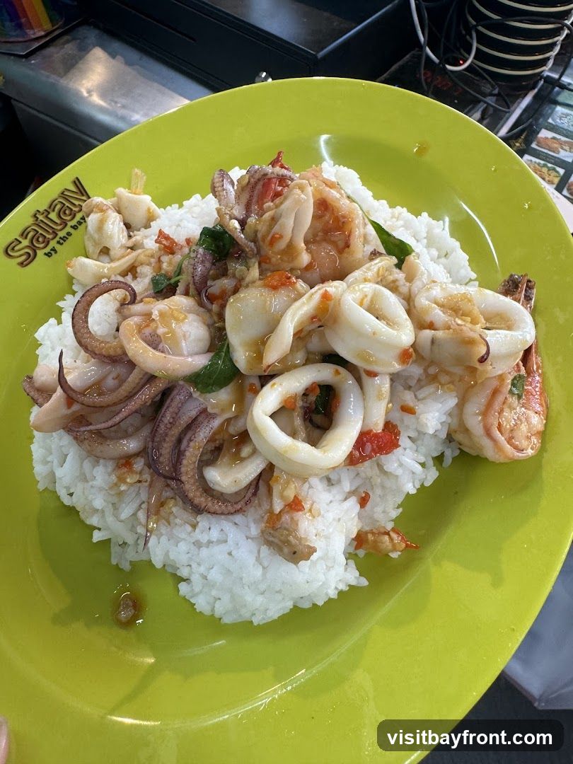 A bright green plate filled with white rice topped with a stir-fry of squid rings, tentacles, and shrimp mixed with chili, basil, and other seasonings. The logo 'Sataw by the bay' is visible on the edge of the plate.