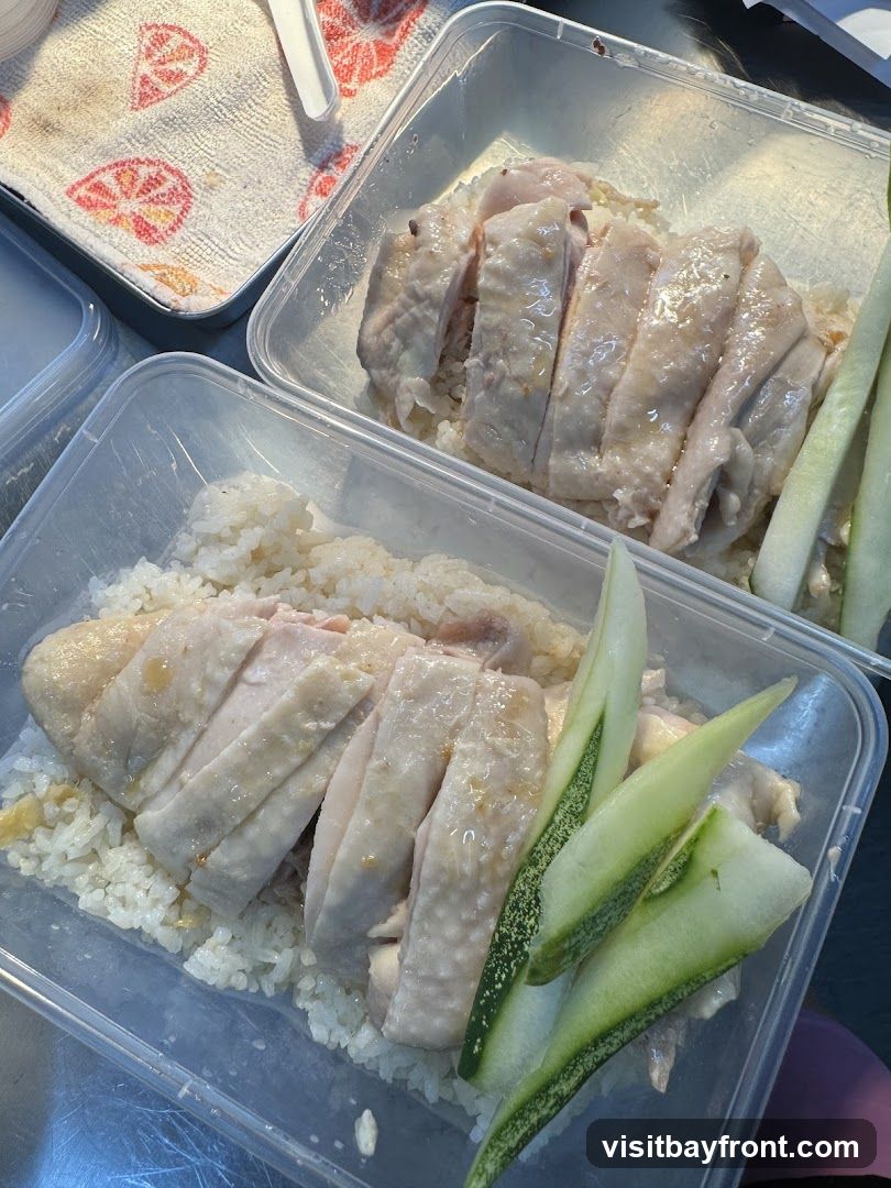 Two takeaway containers filled with Hainanese chicken rice. Each container has a bed of white rice topped with sliced poached chicken and garnished with cucumber slices. A patterned cloth is visible in the upper left corner.