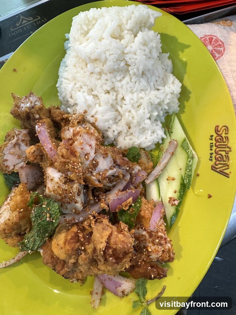 A bright yellow plate holds a generous portion of white rice next to a salad of fried chicken pieces, red onions, mint leaves, and cucumber slices. The chicken is coated in a crispy batter and seasoned with spices. The plate has a logo that reads "Saitay by the bay" and another logo that reads "OOH FU THAI" is partially visible in the upper left corner.