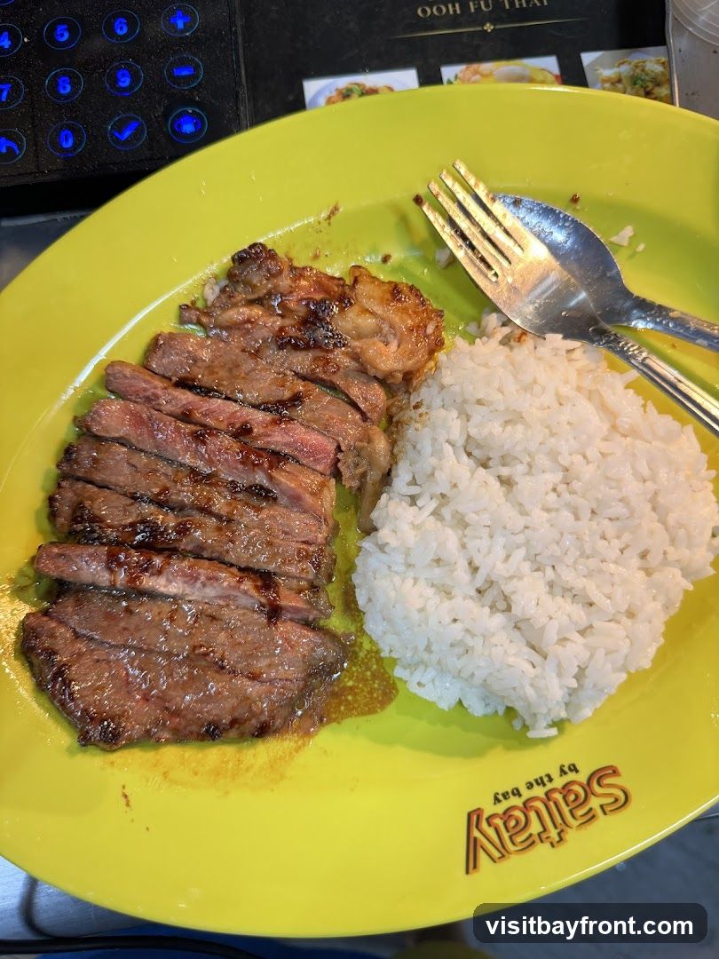A plate of sliced steak with a side of white rice, served with a fork and spoon. The plate is yellow and has a logo that says "Satay by the bay". In the background, there is a menu board with the text "OOH FU THAI" and images of food.