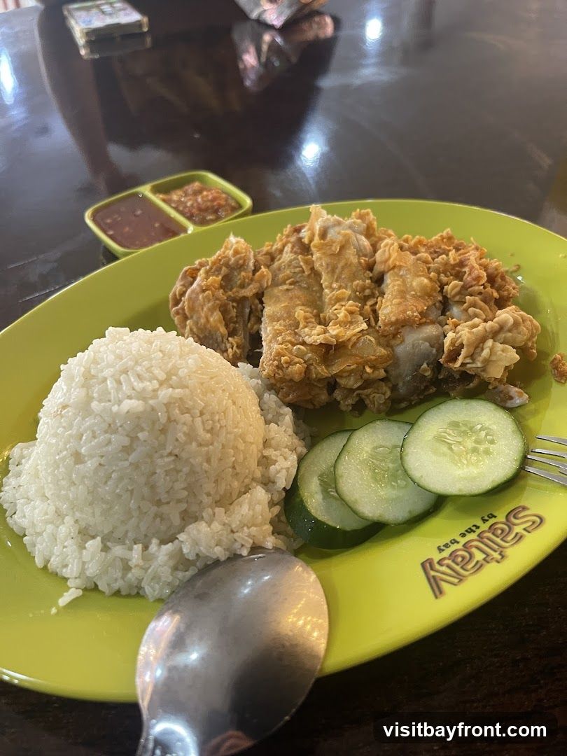 A plate of fried chicken served with a mound of white rice and sliced cucumbers. Two small dishes of dipping sauce are visible in the background. The plate has the logo "Saitray by the bay" printed on it.
