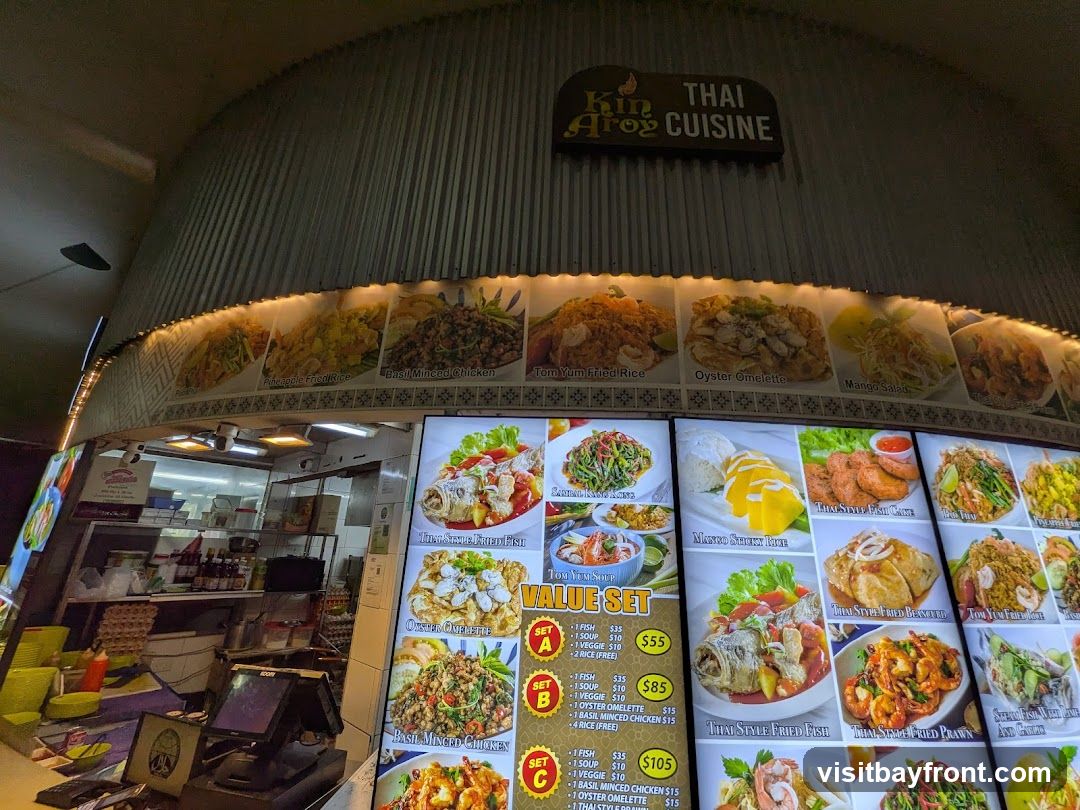 A food stall display featuring a variety of Thai dishes with images and prices. The stall's name, 'Kin Aroy Thai Cuisine', is visible above the display. The menu includes options like Tom Yum Fried Rice, Oyster Omelette, Mango Salad, and various value sets.