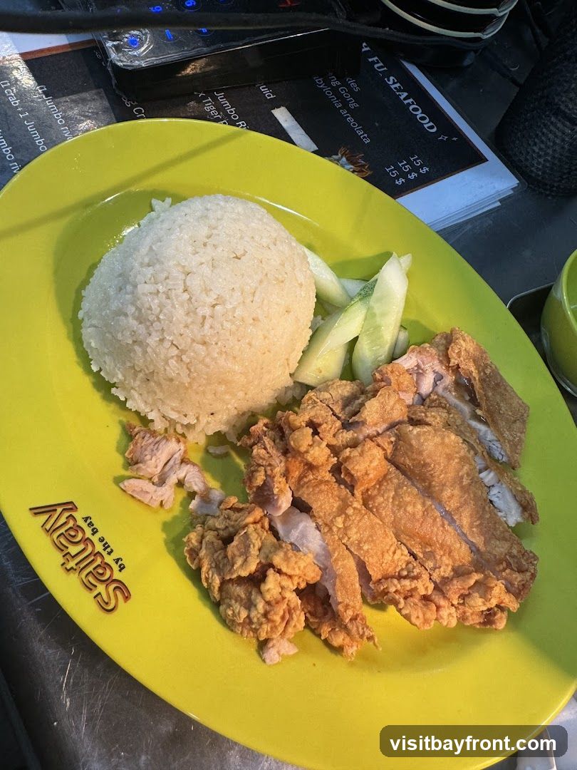 A plate of fried chicken with rice and cucumber slices is shown. The plate is bright yellow and has the "Satay by the bay" logo. A menu is partially visible in the background.