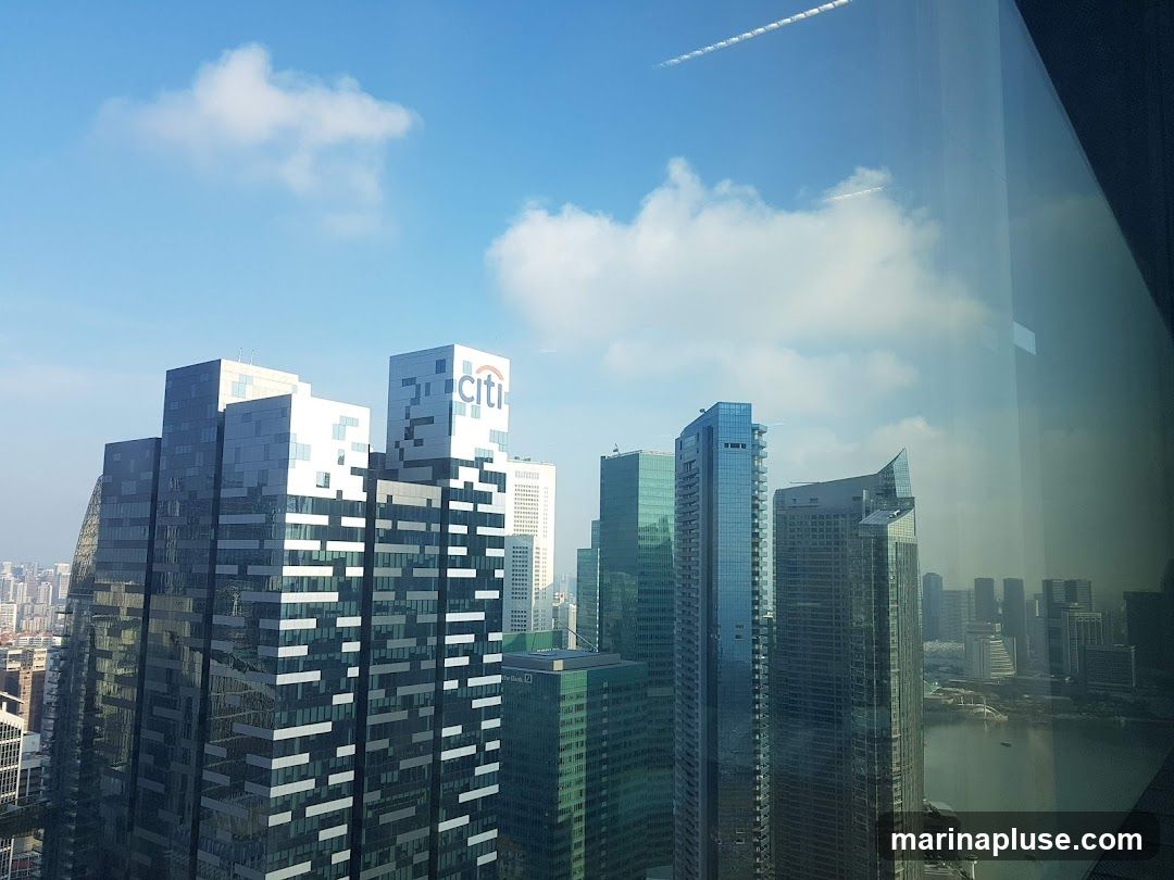 A view of a modern city skyline featuring several tall buildings, including one with the 'citi' logo prominently displayed. The buildings are a mix of glass and metallic facades, reflecting the blue sky and scattered clouds. In the distance, a body of water with a few boats can be seen, along with more urban development. The image appears to be taken from a high vantage point, possibly through a window, as indicated by reflections on the right side.
