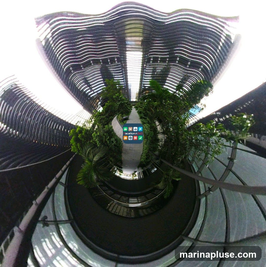 A panoramic view of a modern building's interior, featuring a walkway lined with lush green plants and trees. The architecture is characterized by curved, layered structures with a metallic sheen, creating a futuristic and airy atmosphere. The walkway curves through the space, leading the eye towards a bright, open area at the end. A small cluster of app icons is superimposed on the walkway, with the text "acation n360" visible.