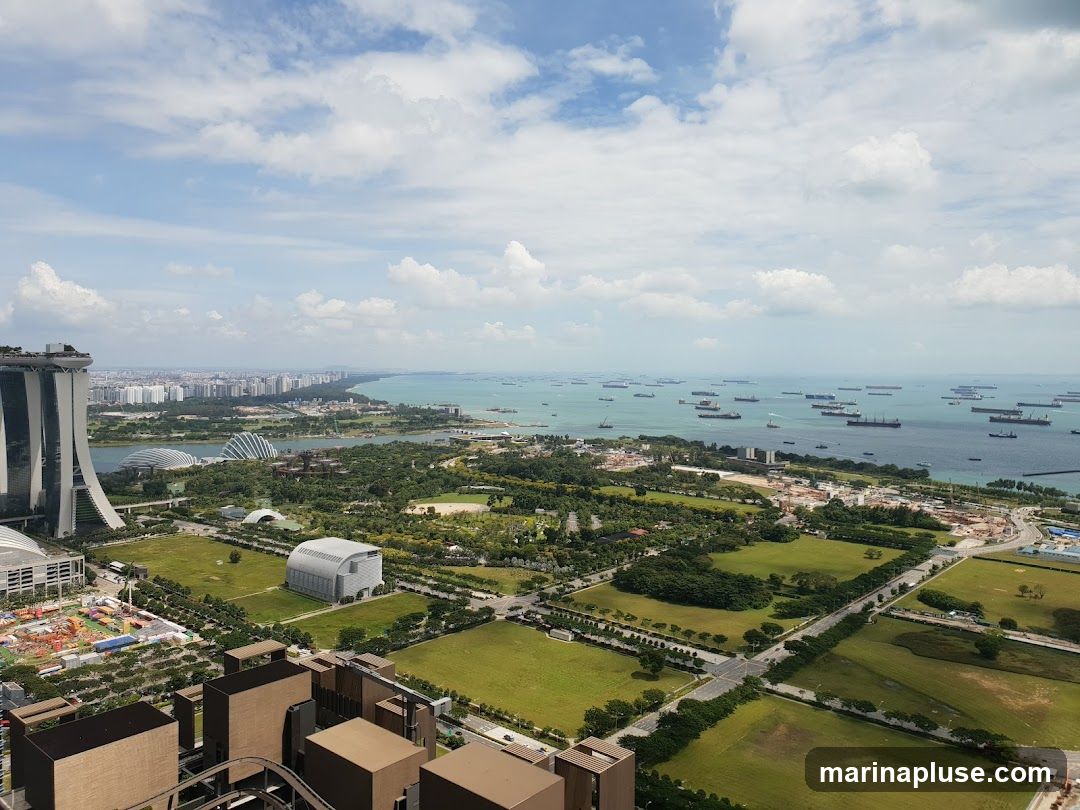 An aerial view captures the expansive Singapore skyline, featuring the iconic Marina Bay Sands hotel on the left. Beyond it, the Gardens by the Bay with its distinctive domes is visible. The foreground shows urban development with buildings and green spaces, while the background is dominated by the vast ocean filled with numerous cargo ships, indicating a busy port. The sky is partly cloudy with a bright, sunny atmosphere.