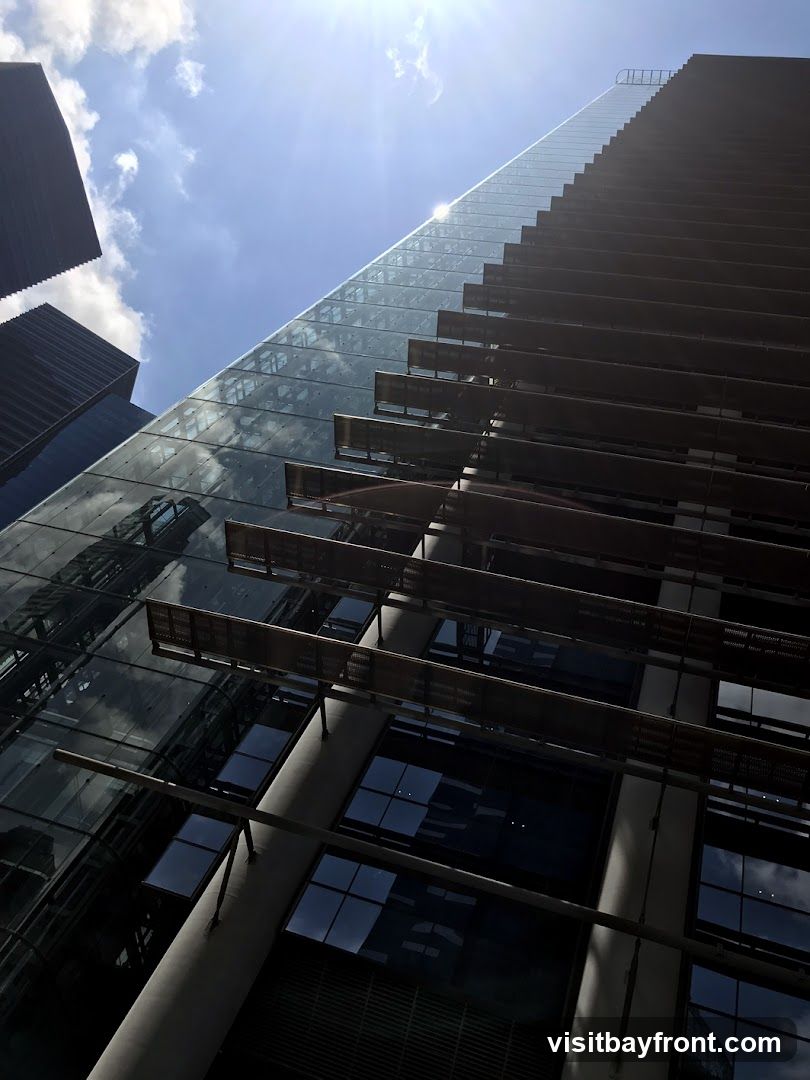 A low-angle shot of a modern skyscraper with a glass facade reflecting the sky and clouds. The building features horizontal louvers or sunshades that create a striped pattern. The sun is shining brightly, creating lens flare and casting shadows. Other buildings are visible in the background.