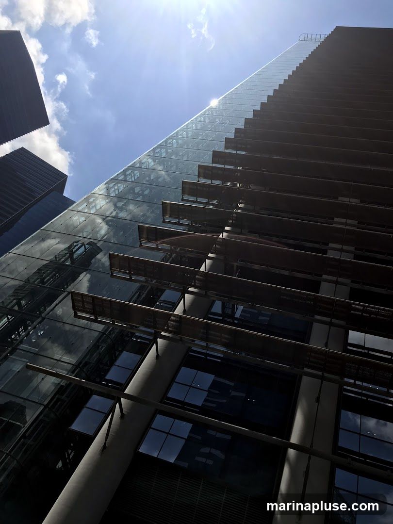 A low-angle shot of a modern skyscraper with a glass facade reflecting the sky and clouds. The building features horizontal louvers or sunshades that create a striped pattern. The sun is shining brightly, creating lens flare and casting shadows. Other buildings are visible in the background.