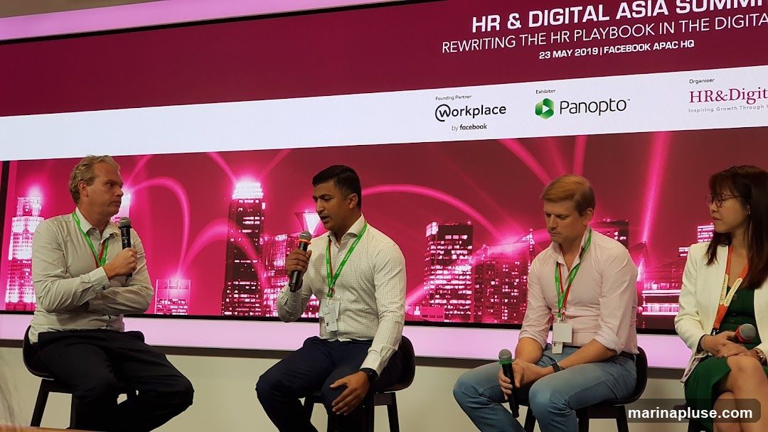 Four people are seated on a stage, participating in a panel discussion. The backdrop features a large screen displaying "HR & DIGITAL ASIA SUMMIT REWRITING THE HR PLAYBOOK IN THE DIGITAL AGE" along with logos for "Workplace by Facebook," "Panopto," and "HR&Digital." The screen also shows a stylized image of city skyscrapers with pink glowing connections. The panelists are holding microphones and appear to be engaged in conversation.