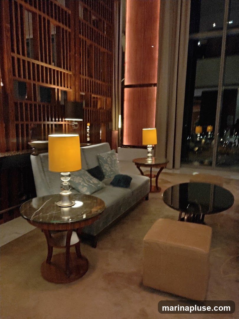 A dimly lit lounge area with a gray sofa, marble-topped side tables, a leather ottoman, and a large wooden lattice wall. Lamps with yellow shades provide warm lighting. Through the window, city lights are visible.
