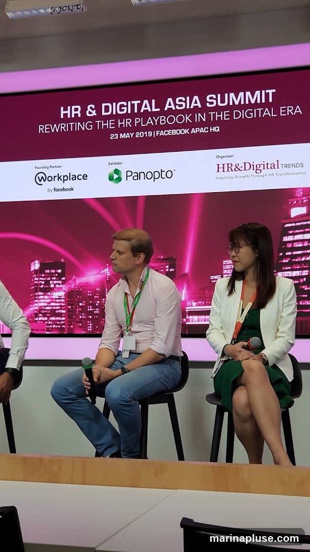 A screen displays the title "HR & DIGITAL ASIA SUMMIT REWRITING THE HR PLAYBOOK IN THE DIGITAL ERA" along with event details and sponsor logos. Two individuals, a man and a woman, are seated on chairs in front of the screen, facing to the side. The man is wearing a light pink shirt and blue jeans, holding a microphone. The woman is wearing a white blazer over a green dress, also holding a microphone. The background features a stylized image of a city skyline with pink lighting.