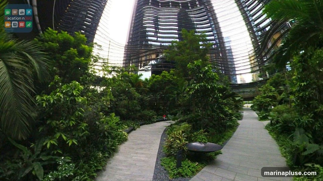 A lush indoor garden with winding pathways and abundant greenery, set against the backdrop of a modern, curved glass and steel building. The scene captures a serene and natural environment within an urban setting.