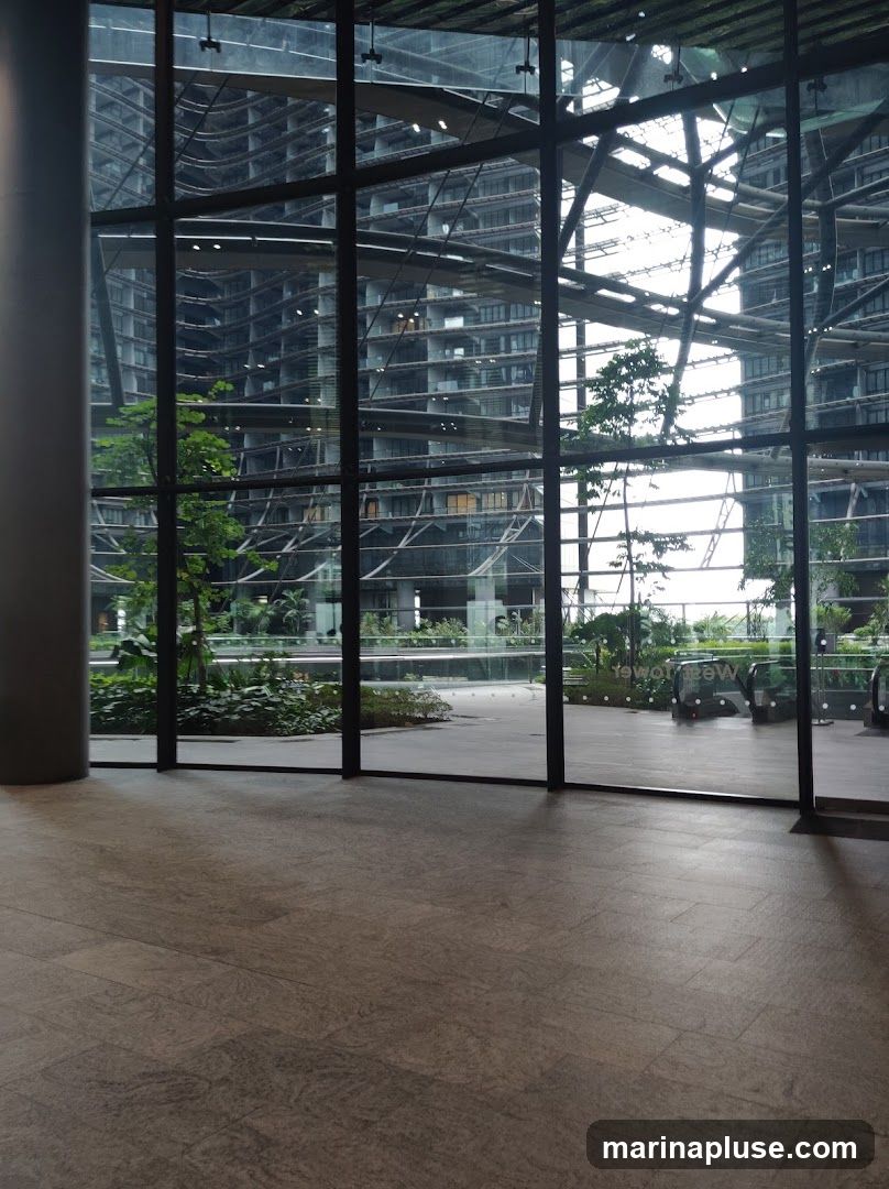 View through a large glass facade of a modern building, revealing a multi-story structure with intricate architectural details and balconies. Lush green plants and trees are visible in the foreground and on the building's terraces, creating a biophilic design. The floor is tiled with large, grey stone-like tiles. In the distance, automated walkways or escalators are partially visible. The overall impression is one of a contemporary, nature-integrated urban space.