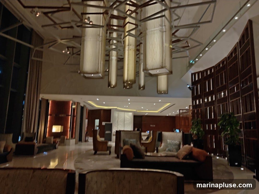 A wide shot of a modern hotel lobby with high ceilings and contemporary decor. Large, rectangular pendant lights hang from the ceiling, casting a warm glow. The lobby features comfortable seating areas with plush sofas and armchairs, arranged around a central reception area. A decorative wooden screen divides parts of the space, adding a touch of elegance. Plants are strategically placed to enhance the ambiance. The overall impression is one of luxury and sophistication.