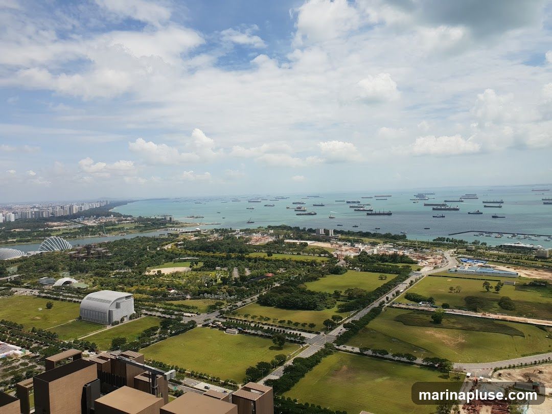 An aerial view captures a sprawling coastal city under a partly cloudy sky. The foreground shows urban development with buildings and green spaces. Further out, a large body of water is filled with numerous cargo ships and smaller vessels, indicating a busy port. The coastline stretches into the distance, lined with more buildings and lush greenery.