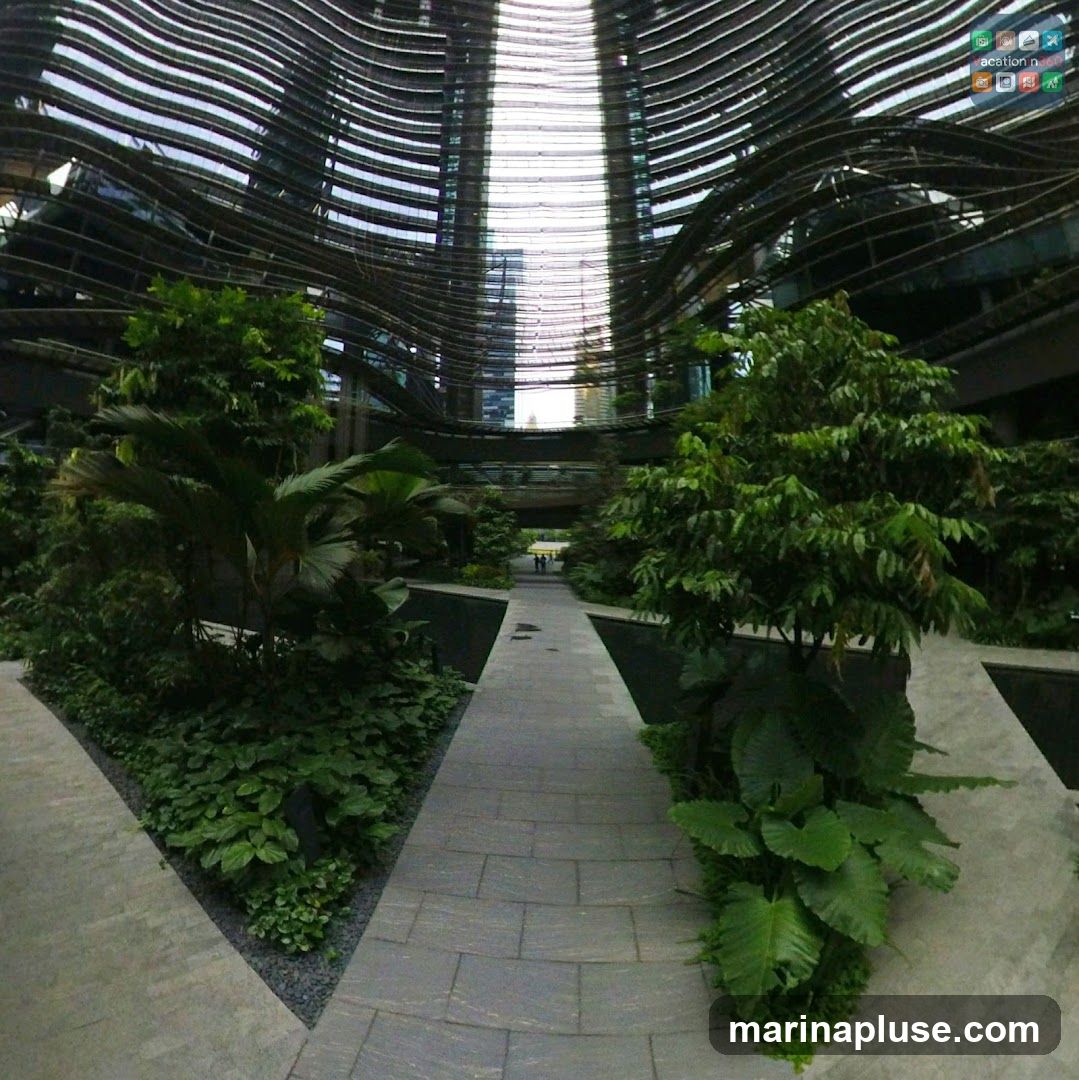 A wide-angle view of a modern architectural space featuring a central walkway lined with lush green plants and water features. The surrounding buildings have a distinctive wavy, layered facade made of dark material and glass, creating a dramatic, enclosed atmosphere. The walkway leads towards a bright, open area in the distance where a few people can be seen.