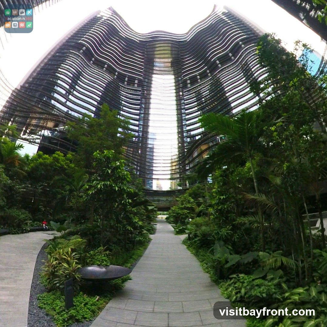 A wide-angle shot captures the striking architecture of a modern building with a curved, wave-like facade composed of horizontal metallic louvers. The building's interior is visible through the gaps, revealing a bright, open space. Lush green trees and tropical plants line a paved walkway that curves through the foreground, creating a serene and natural ambiance within an urban setting. A lone figure in red is visible in the distance on the left path.