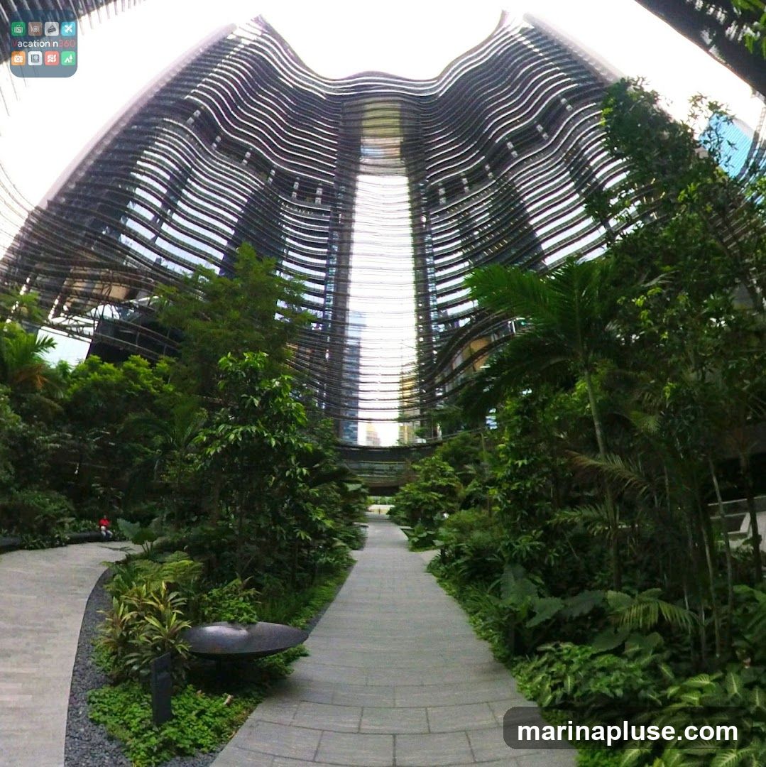 A wide-angle shot captures the striking architecture of a modern building with a curved, wave-like facade composed of horizontal metallic louvers. The building's interior is visible through the gaps, revealing a bright, open space. Lush green trees and tropical plants line a paved walkway that curves through the foreground, creating a serene and natural ambiance within an urban setting. A lone figure in red is visible in the distance on the left path.