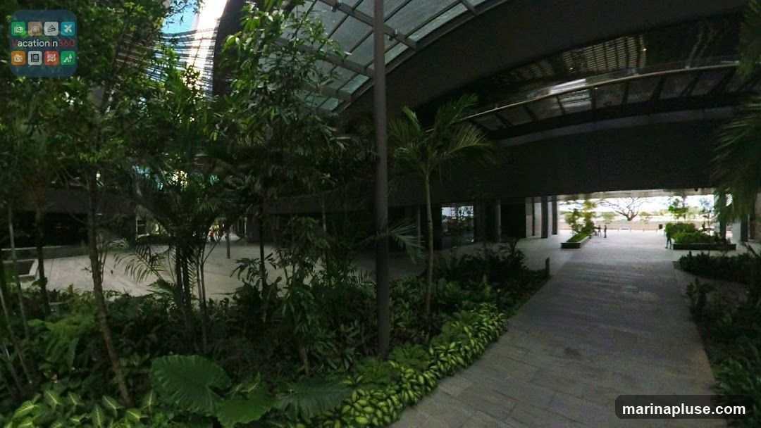 A wide outdoor walkway paved with large stone tiles is lined with abundant tropical plants and trees. The walkway is covered by a modern, sloped glass and metal roof structure, allowing natural light to filter through. In the distance, the walkway opens up to a brighter outdoor area with more greenery and a large tree. A few people are visible in the distance, suggesting a public space or commercial area. A small logo with the text "Vacation n360" and several icons is present in the top left corner of the image.