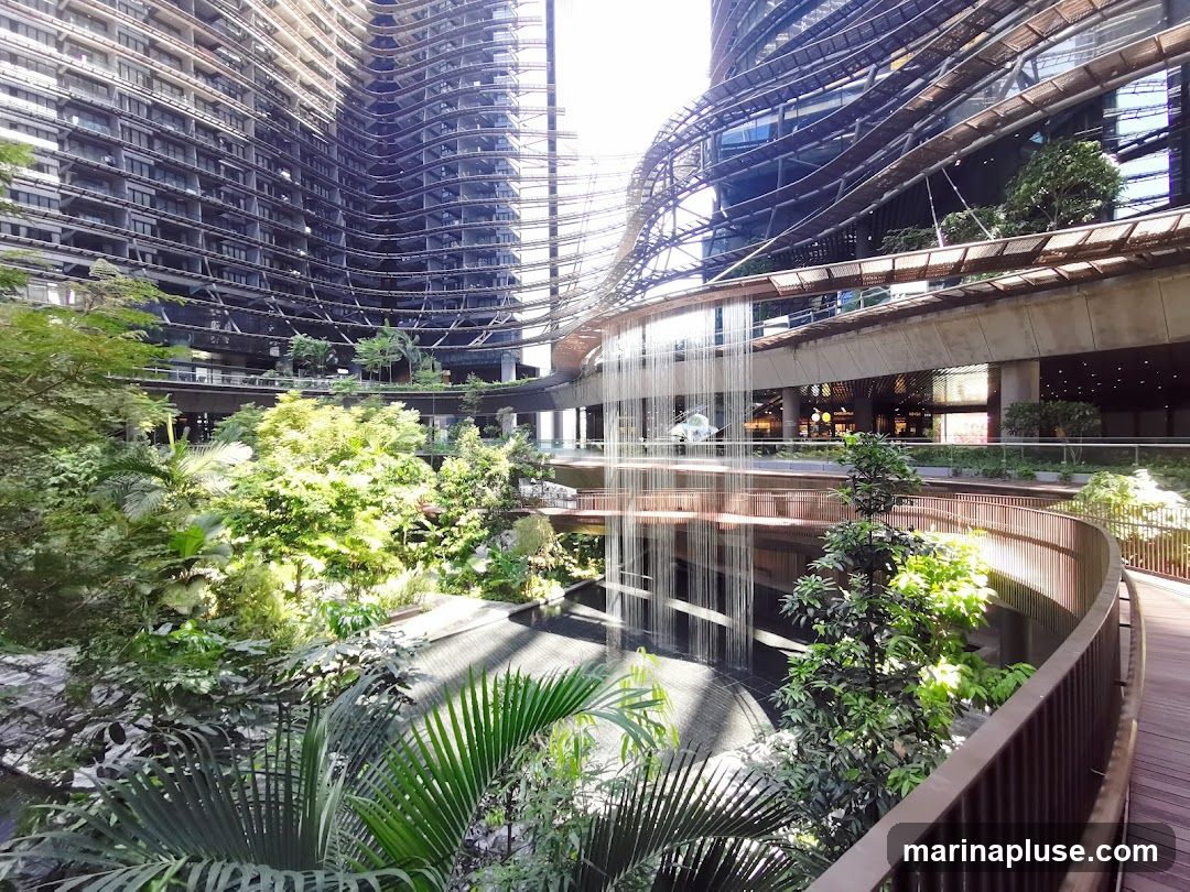 A view of a multi-level atrium with a large indoor waterfall cascading down. Lush green plants and trees are abundant throughout the space, creating a natural oasis within a modern architectural setting. A wooden walkway with a railing curves through the foreground, offering a vantage point to observe the scene. The background features the curved facades of tall buildings with intricate structural details and glass windows. Several shops are visible on a lower level, illuminated by interior lighting.