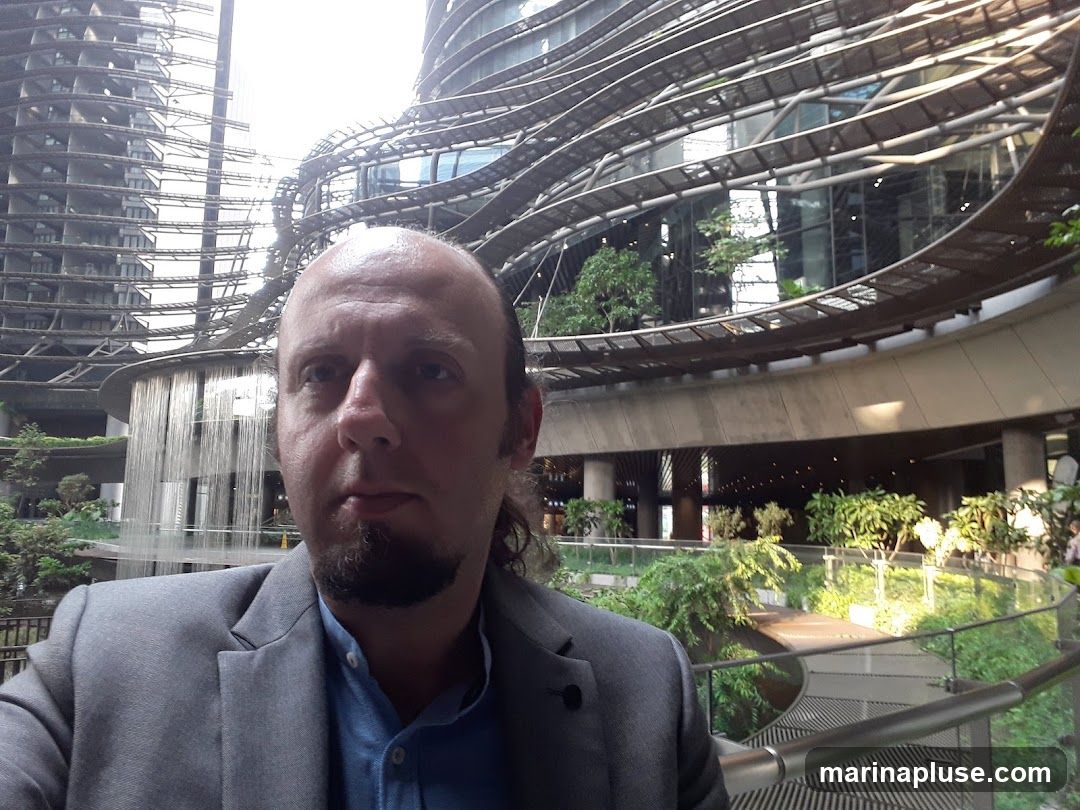 A man in a grey suit and blue shirt takes a selfie in front of a striking modern building with curved walkways and greenery. The building features a unique, layered design with exposed structural elements and large glass windows. Water cascades down a circular structure in the background, adding to the dynamic visual. The overall scene suggests an urban environment with a focus on contemporary architecture and landscaping.