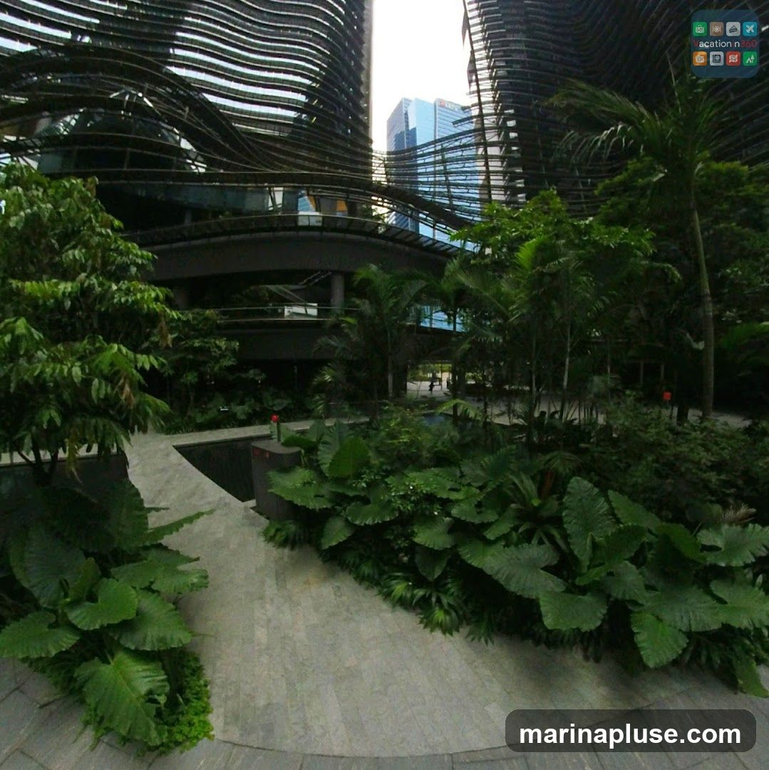 A view of contemporary buildings with wavy facades, surrounded by abundant tropical plants and trees. A paved walkway curves through the greenery, leading towards the structures. In the background, a blue skyscraper stands tall. The scene suggests a blend of urban design and nature.