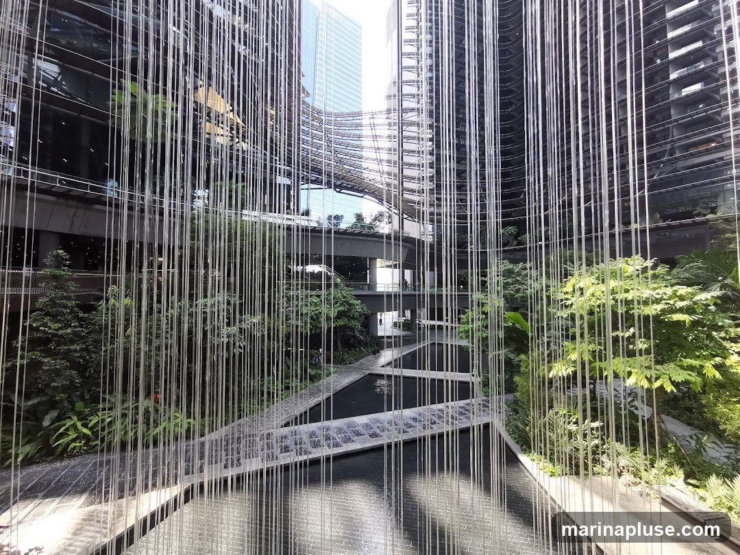 A view through a curtain of vertical metallic strands into a modern courtyard. The courtyard features tiered water channels and lush greenery, with contemporary architecture forming the backdrop. Sunlight reflects off the metallic strands and the water, creating a dynamic interplay of light and shadow.