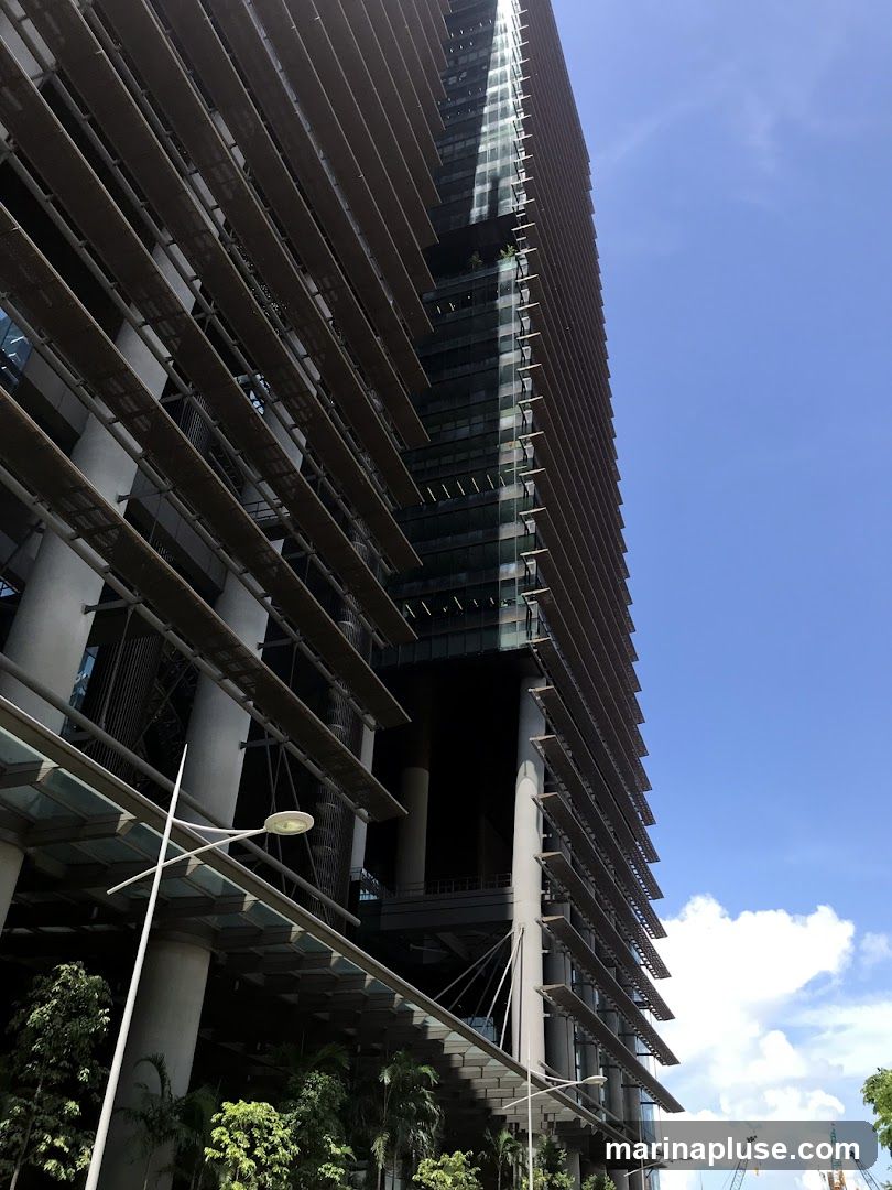 A low-angle shot of a modern skyscraper with a distinctive facade featuring horizontal louvers. The building has a dark glass exterior and is supported by large, white columns. The sky is bright blue with scattered white clouds. Trees and greenery are visible at the base of the building. Construction cranes can be seen in the distance.