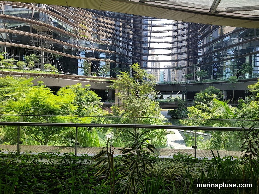 A view into a multi-level interior courtyard filled with abundant green plants and trees. The architecture features curved glass facades and walkways, with scaffolding visible on one side, suggesting ongoing construction or maintenance. The overall impression is a blend of modern design and natural elements.