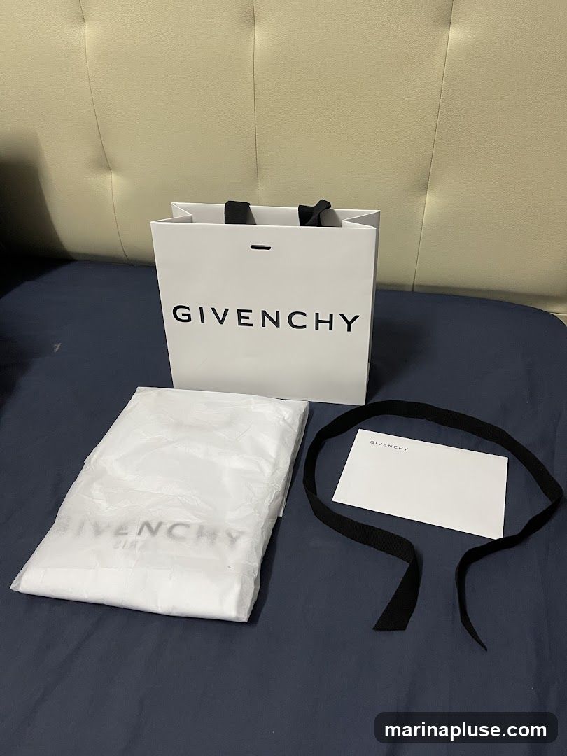 A white Givenchy shopping bag with black handles is placed on a dark blue surface. Next to it is a white dust bag, also with the Givenchy logo faintly visible, and a small white card with the Givenchy logo and a black ribbon. The items are arranged on a surface that appears to be a bed with a tufted headboard in the background.