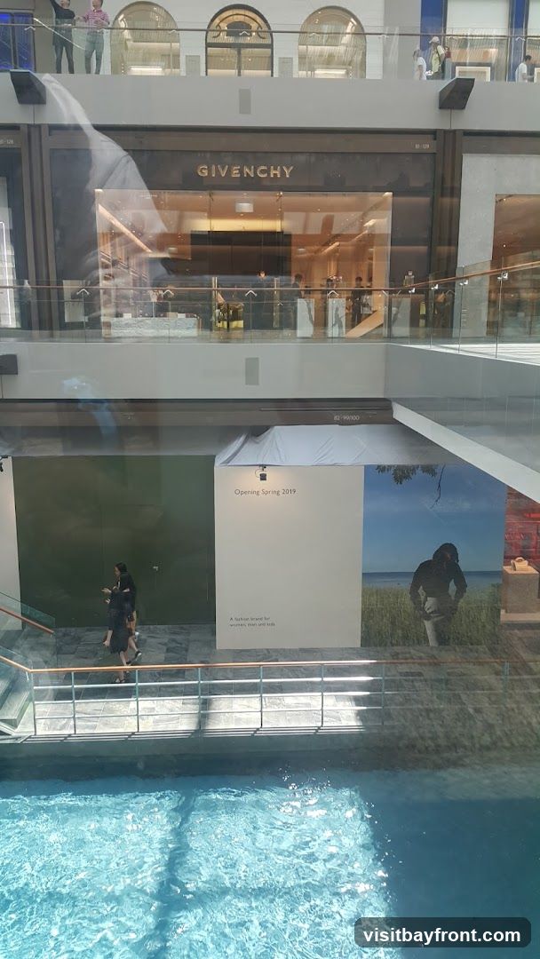 A view of a Givenchy store with its name prominently displayed above the entrance. Inside, mannequins and displays are visible. Below the store, a large advertisement for an upcoming opening in Spring 2019 is displayed, featuring text that reads "A fashion brand for women, men and kids" and a scenic image. A body of water is visible in the foreground.