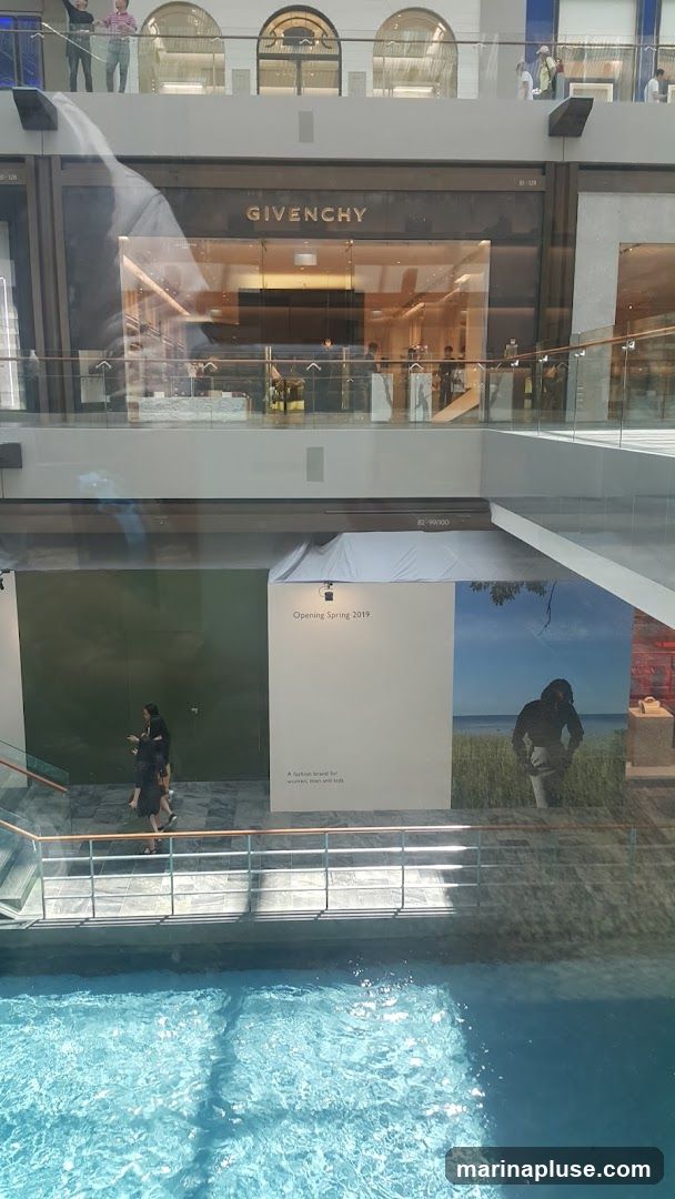 A view of a Givenchy store with its name prominently displayed above the entrance. Inside, mannequins and displays are visible. Below the store, a large advertisement for an upcoming opening in Spring 2019 is displayed, featuring text that reads "A fashion brand for women, men and kids" and a scenic image. A body of water is visible in the foreground.