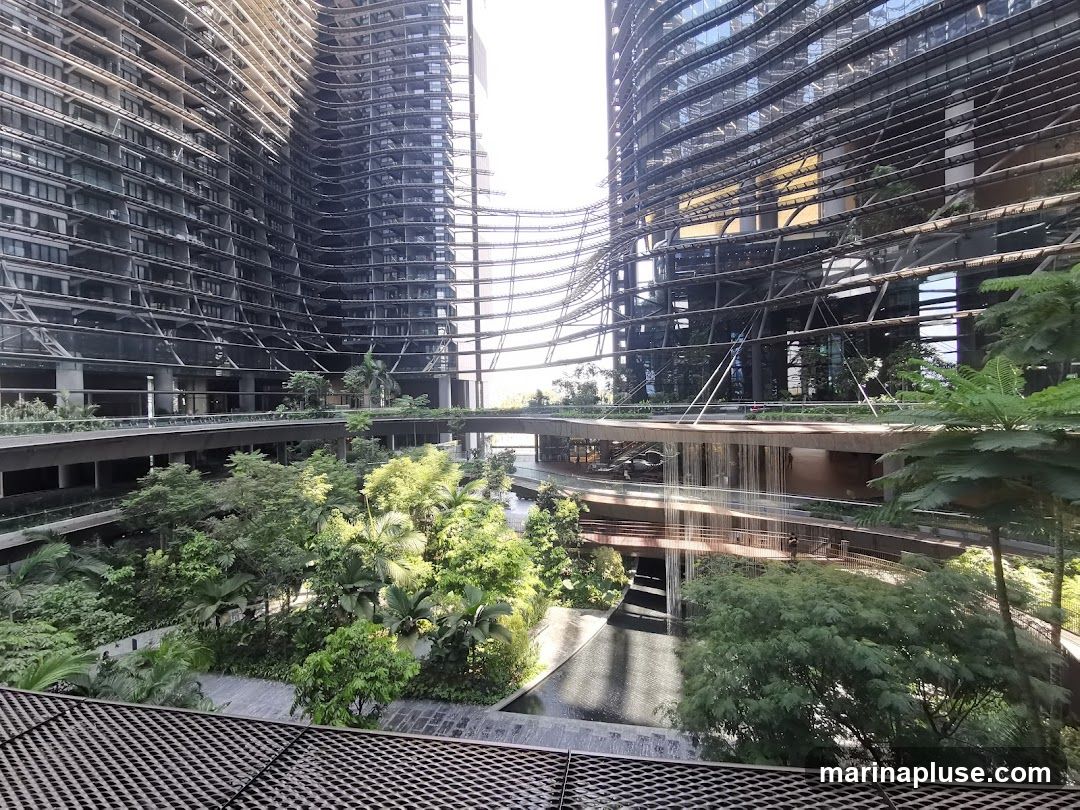 A view of a modern architectural complex with curved glass facades and multiple levels of walkways and balconies. Lush green trees and plants are integrated into the design, creating a striking contrast between the man-made structures and nature. Water features are visible, adding to the serene atmosphere.