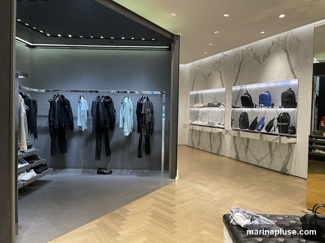 The interior of a modern, upscale store displays men's clothing and accessories. Racks of black and light blue jackets, shirts, and pants are visible on the left. Shelves on the right showcase sneakers and various backpacks and bags, some with the Givenchy logo. The store features marble-patterned walls and a herringbone wood floor.