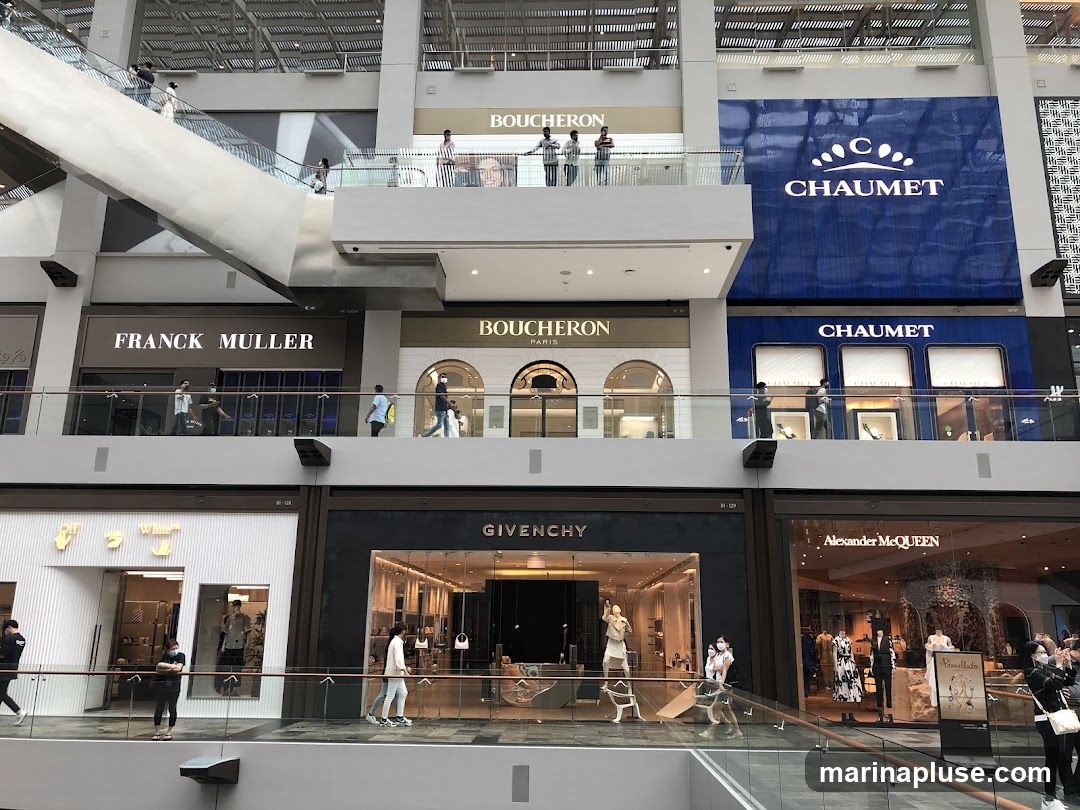 A multi-level interior view of a modern shopping mall featuring several high-end luxury brand stores. Visible storefronts include Franck Muller, Boucheron, Chaumet, Off-White, Givenchy, and Alexander McQueen. People are seen walking on the elevated walkways and inside the stores.