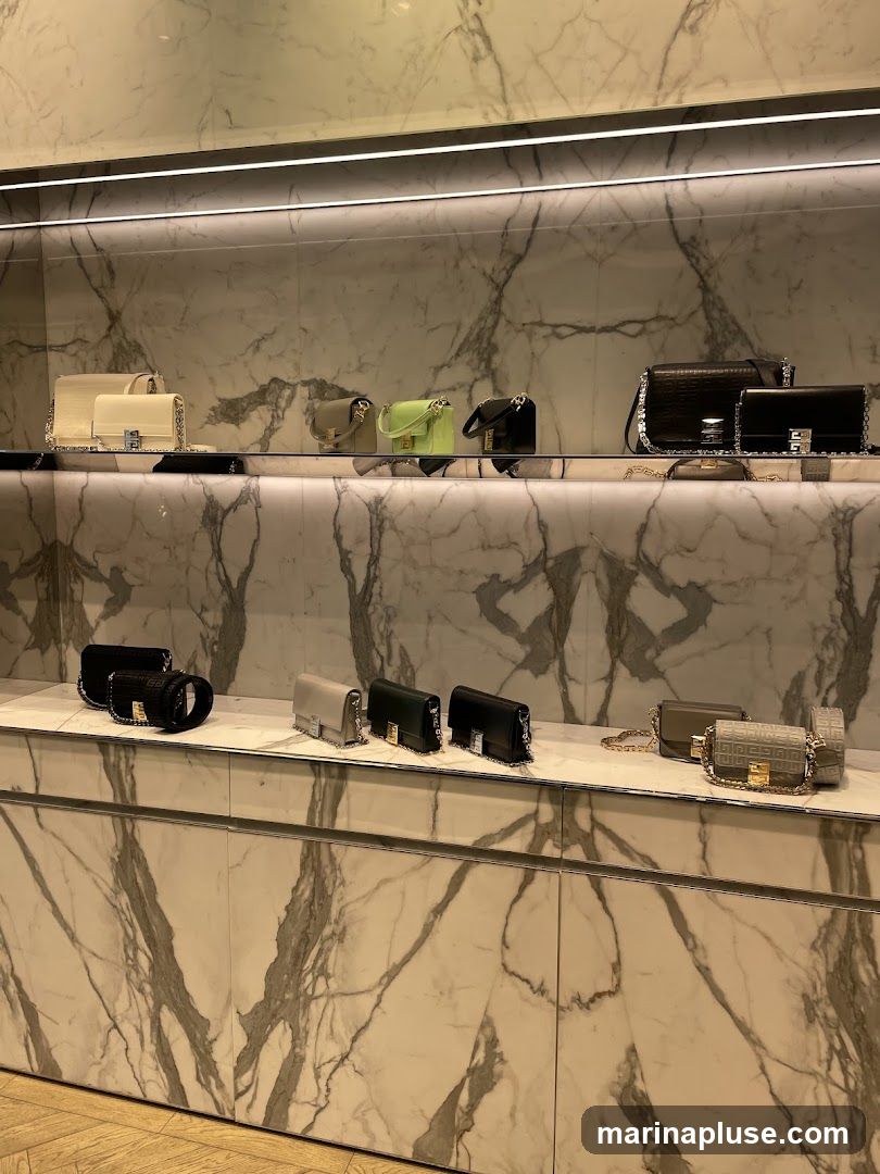 A display of various handbags on shelves with a marble-patterned background. The handbags are in different colors including cream, lime green, black, and grey. Some have chain straps and decorative clasps.