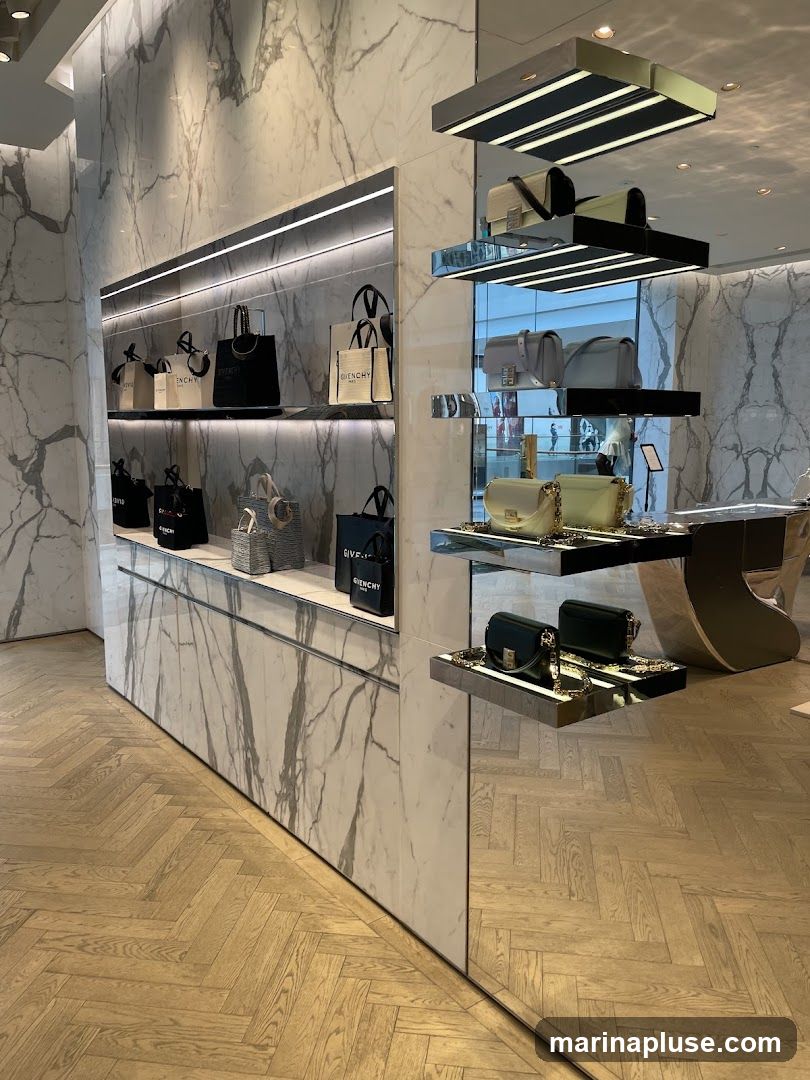 A well-lit interior display of various handbags on shelves. The walls are adorned with marble-patterned panels, and the floor is laid with light-colored wooden planks in a herringbone pattern. Several handbags, some with the Givenchy logo visible, are showcased on sleek, modern shelving units. The overall aesthetic is one of luxury and sophistication.