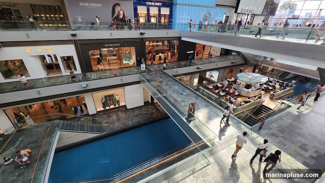 An aerial view of the interior of a modern shopping mall. Multiple levels are visible, connected by escalators and walkways. Various luxury brand stores like Boucheron, Chaumet, Hublot, Chanel, Givenchy, and Off-White are identifiable by their signage. A central atrium features a canal with a boat, and a cafe with outdoor seating. Shoppers are seen walking on the different levels and bridges.