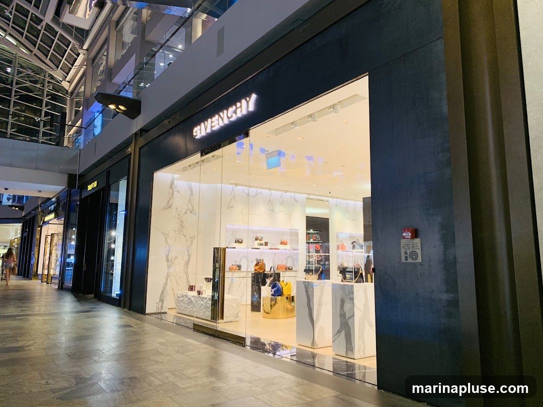 The exterior of a Givenchy store with its name illuminated above the entrance. The store features a modern design with large glass windows displaying handbags and accessories on marble pedestals. The interior is brightly lit, showcasing a selection of luxury goods. To the right of the entrance, a "HOSE REEL" sign is visible on the dark facade.
