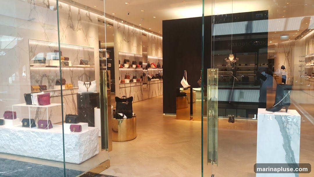 A view through a glass storefront into a brightly lit, modern luxury store. The interior features marble-patterned walls and displays showcasing a variety of handbags and shoes. Several handbags are arranged on tiered white and black marble display stands in the foreground. In the background, shelves are filled with more handbags. To the right, a black boot with a silver accent is displayed on a marble pedestal. A person in a white top and dark pants can be seen in the background, possibly a sales associate. The store appears to be well-organized and upscale.