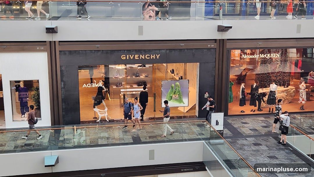 A wide shot of the Givenchy store entrance in a mall. The store has a dark facade with the gold 'GIVENCHY' logo prominently displayed above the entrance. The large glass windows showcase handbags and mannequins dressed in clothing. Several people are walking past the store. To the right of the Givenchy store is an Alexander McQueen store with a similar display of mannequins and merchandise.