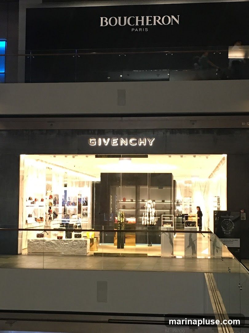 The image shows the brightly lit storefront of a luxury brand, Givenchy, located in a mall. The store displays handbags and accessories on shelves and mannequins. Above the Givenchy store, the Boucheron Paris logo is visible on a darker facade.